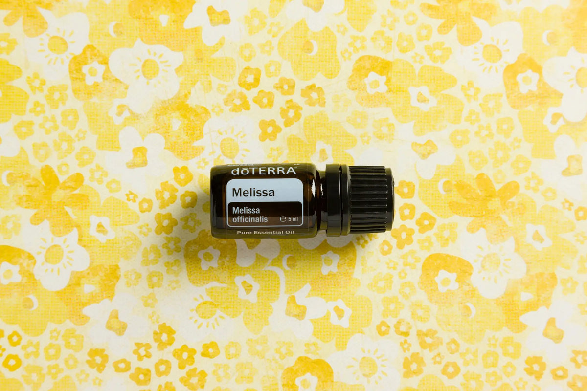 Aceite esencial de Melissa dōTERRA 5ml sobre tela estampada floral en tonos amarillos y blancos, flat lay