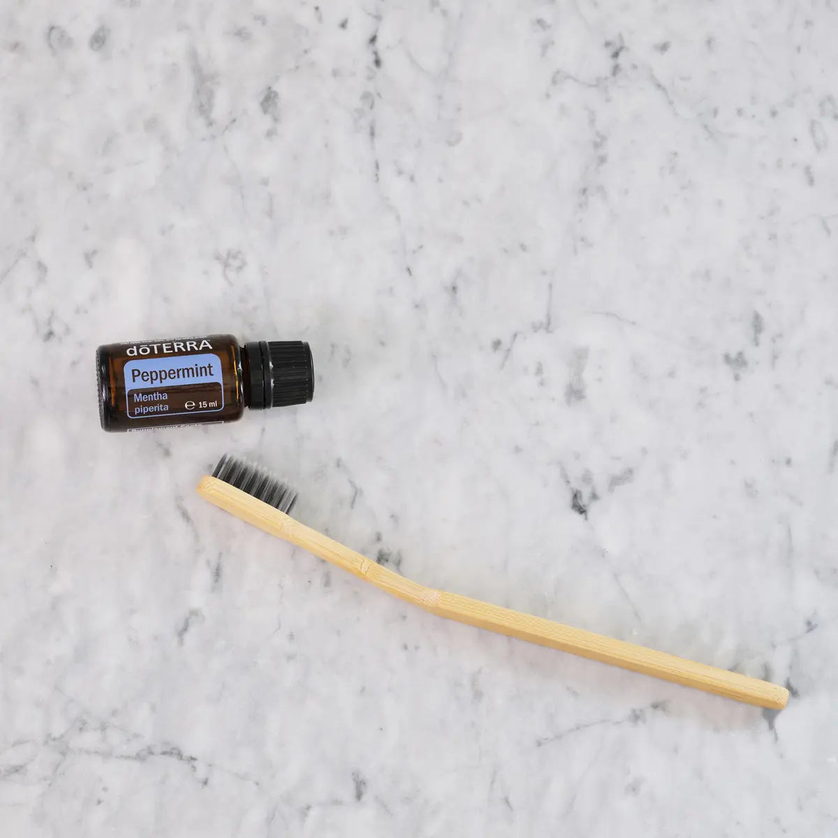 Aceite esencial Peppermint dōTERRA 15ml junto a cepillo de dientes de bambú sobre mármol blanco, vista cenital