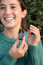 Mujer joven con pelo castaño recogido sonriendo sosteniendo frasco de aceite esencial Peppermint dōTERRA 15ml abierto, jersey verde turquesa, exterior