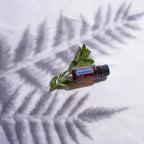 Aceite esencial Peppermint dōTERRA 15ml junto a ramita de menta sobre tela blanca con sombra de helecho proyectada