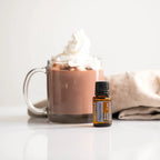 Aceite esencial Peppermint dōTERRA 15ml junto a taza de vidrio con chocolate caliente y nata montada, servilleta de lino, fondo blanco — variante