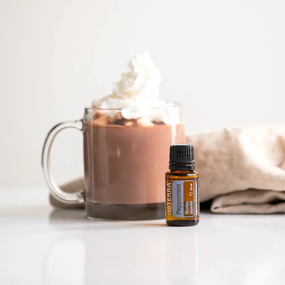 Aceite esencial Peppermint dōTERRA 15ml junto a taza de vidrio con chocolate caliente y nata montada, servilleta de lino, fondo blanco