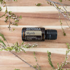 Aceite esencial de Mirra dōTERRA 15ml horizontal sobre tabla de madera cálida rodeado de ramas con flores blancas diminutas, flat lay