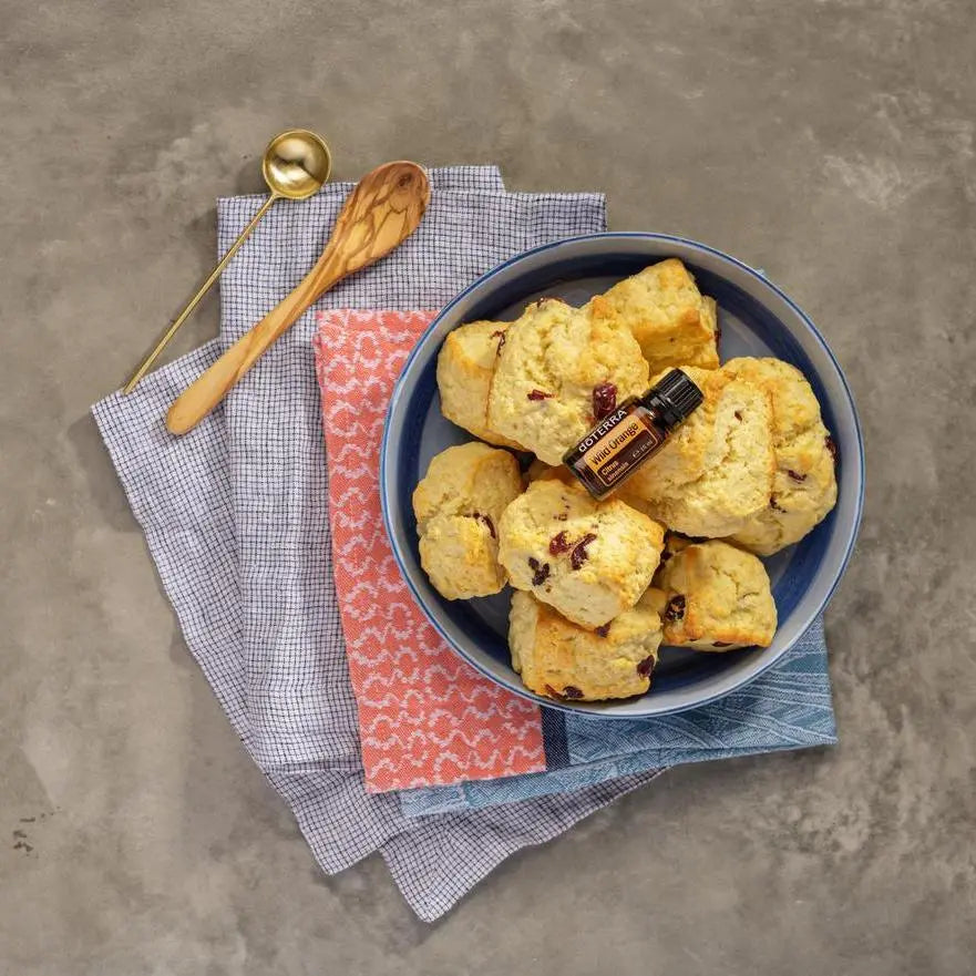 Flat lay del Aceite Esencial de Naranja Silvestre dōTERRA 15ml en cuenco azul lleno de scones con arándanos, servilletas de tela a cuadros y estampada y cucharas de madera y dorada, superficie de piedra gris