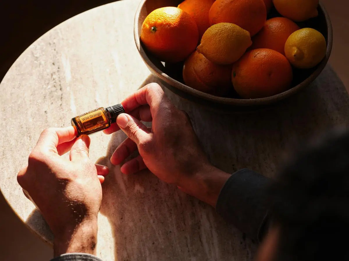 Vista aérea de manos sobre mesa redonda de piedra sosteniendo el Aceite Esencial de Naranja Silvestre dōTERRA 15ml, cuenco de naranjas y limones enteros al fondo, luz cálida dramática