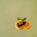 Frasco del Aceite Esencial de Naranja Silvestre dōTERRA 15ml recostado sobre dos mitades de naranja fresca con hojas verdes, fondo verde salvia