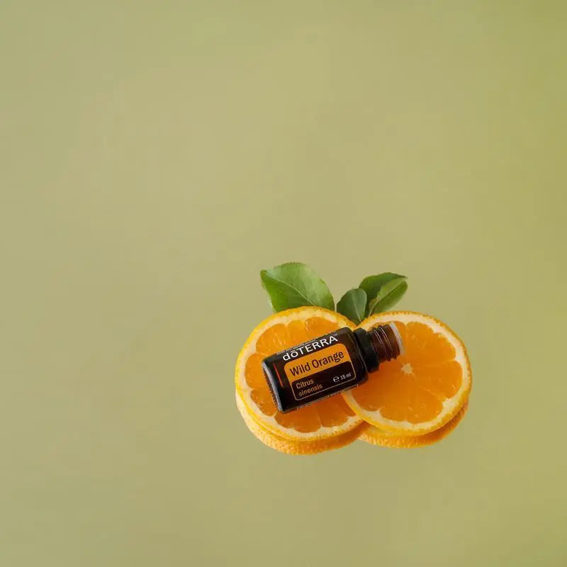 Frasco del Aceite Esencial de Naranja Silvestre dōTERRA 15ml recostado sobre dos mitades de naranja fresca con hojas verdes, fondo verde salvia