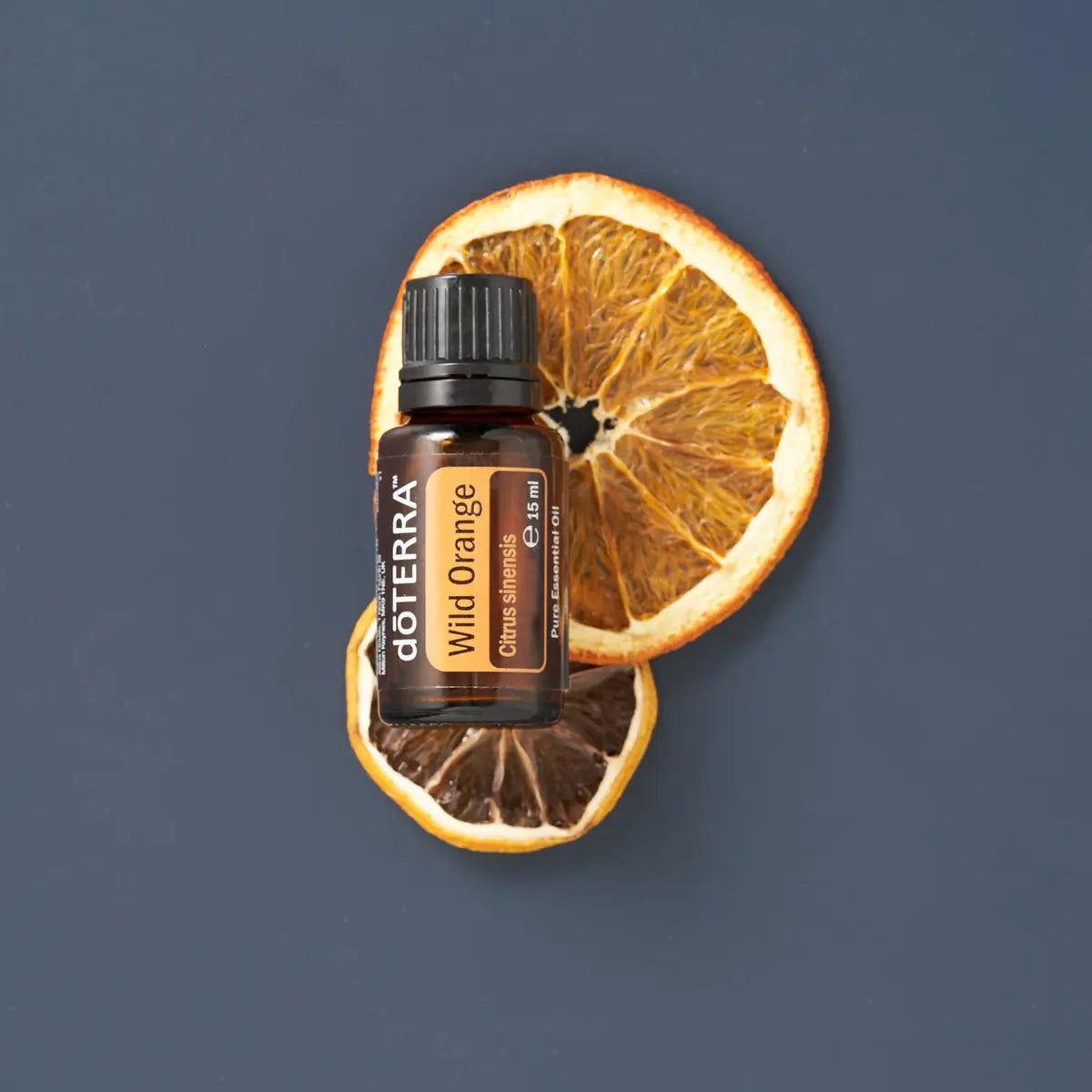 Frasco del Aceite Esencial de Naranja Silvestre dōTERRA 15ml sobre dos rodajas de naranja deshidratada superpuestas, fondo gris azulado