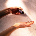 Dos manos sosteniendo frasco de aceite esencial de Pachulí dōTERRA 15ml sobre palma, haz de luz solar y sombras geométricas en pared rosa