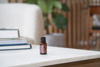 Aceite esencial de Pachulí dōTERRA 15ml sobre mesa de mármol blanca con pila de libros y maceta, estantería de lamas de madera al fondo