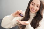 Mujer joven de pelo largo moreno aplicando gota de aceite esencial de Pachulí dōTERRA 15ml en su palma, jersey crema cuello alto, fondo gris