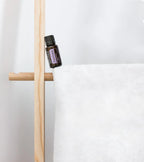 Aceite esencial de Pachulí dōTERRA 15ml apoyado en toallero de madera con toalla de lino blanco, pared blanca al fondo