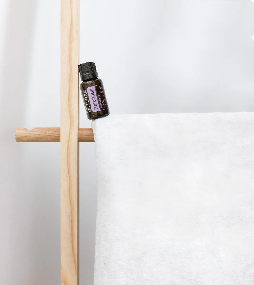 Aceite esencial de Pachulí dōTERRA 15ml apoyado en toallero de madera con toalla de lino blanco, pared blanca al fondo