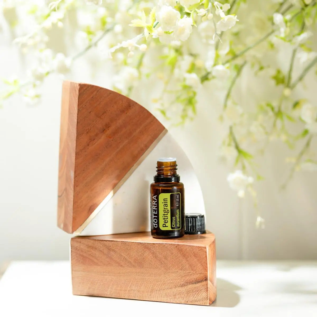 Aceite Esencial Petitgrain dōTERRA 15ml en exhibidor de madera con flores blancas y bokeh suave
