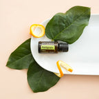Aceite Esencial Petitgrain dōTERRA 15ml en plato blanco con hojas tropicales y piel de naranja cenital