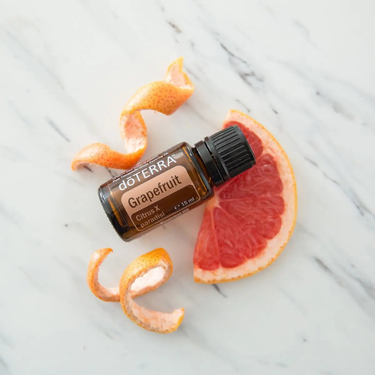Aceite esencial de pomelo dōTERRA 15ml sobre mármol blanco con rodaja de pomelo rojo y espiral de piel cítrica fresca, flat lay cenital