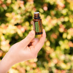 Mano sujetando el Aceite Esencial de Romero dōTERRA 15ml ante fondo bokeh dorado y verde de jardín otoñal, tapa extraída
