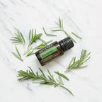 Flat lay del Aceite Esencial de Romero dōTERRA 15ml rodeado de ramas sueltas de romero fresco sobre mármol blanco
