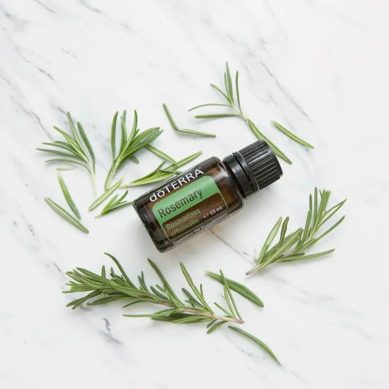 Flat lay del Aceite Esencial de Romero dōTERRA 15ml rodeado de ramas sueltas de romero fresco sobre mármol blanco