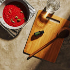 Vista aérea del Aceite Esencial de Romero dōTERRA 15ml sobre tabla de bambú con cuchara de madera, sartén con salsa de tomate y hojas de albahaca a un lado, vaso de agua, superficie de piedra con sombras dramáticas