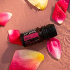 Frasco de aceite esencial de Rosa dōTERRA 5ml recostado en agua rosada con pétalos multicolores y gotas de agua, vista cenital