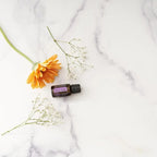 Flat lay del Aceite Esencial de Salvia Romana dōTERRA 15ml sobre mármol blanco con gerbera naranja y ramas de flores blancas de gypsophila