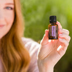 Mujer pelirroja en exterior sosteniendo el Aceite Esencial de Salvia Romana dōTERRA 15ml hacia la cámara, jardín verde desenfocado