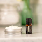 Aceite esencial de Sándalo Hawaiano dōTERRA 5ml junto a tarro de crema de aluminio abierto sobre encimera, planta verde desenfocada al fondo