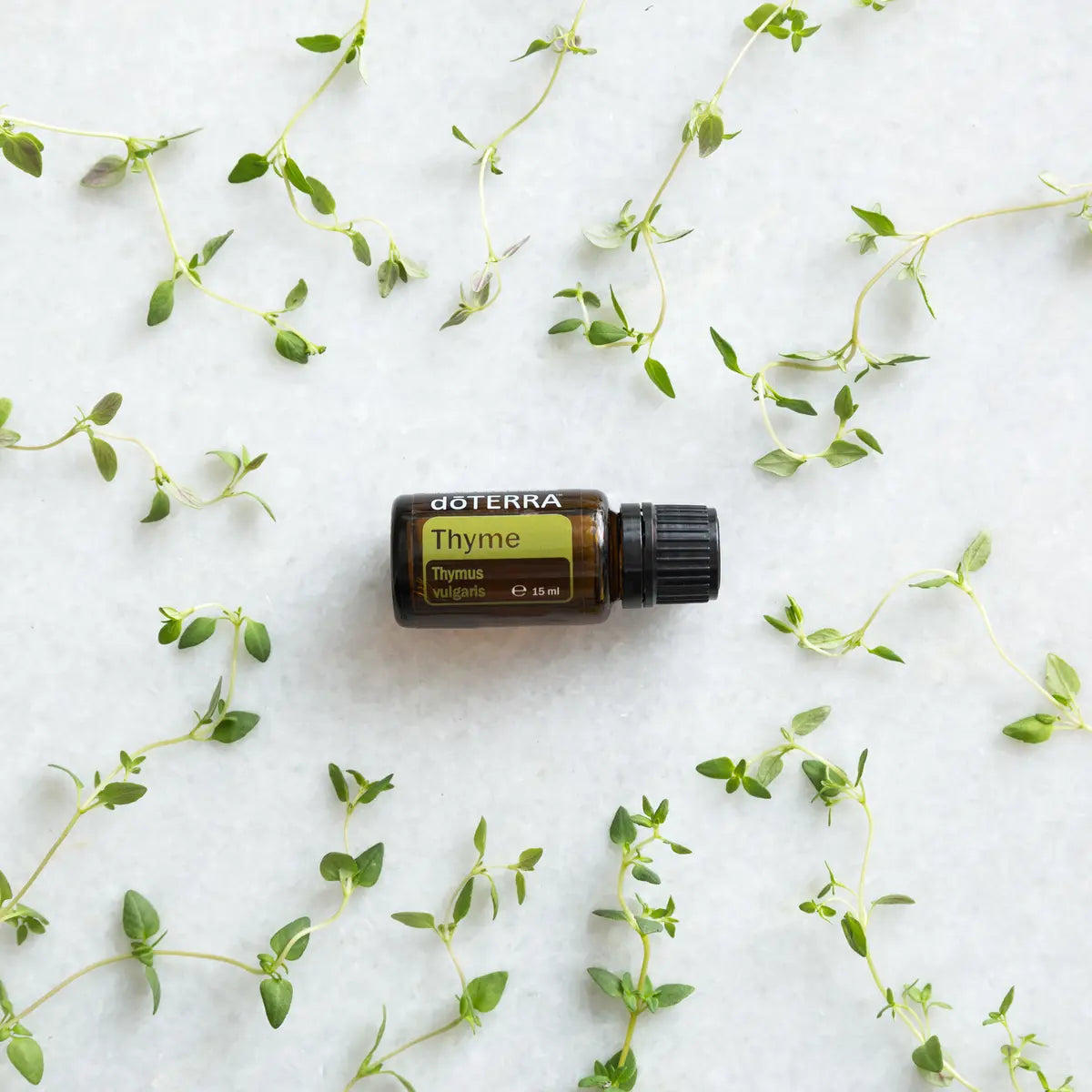 Aceite esencial de Tomillo dōTERRA 15ml rodeado de ramas de tomillo fresco esparcidas sobre superficie de mármol blanco, vista cenital