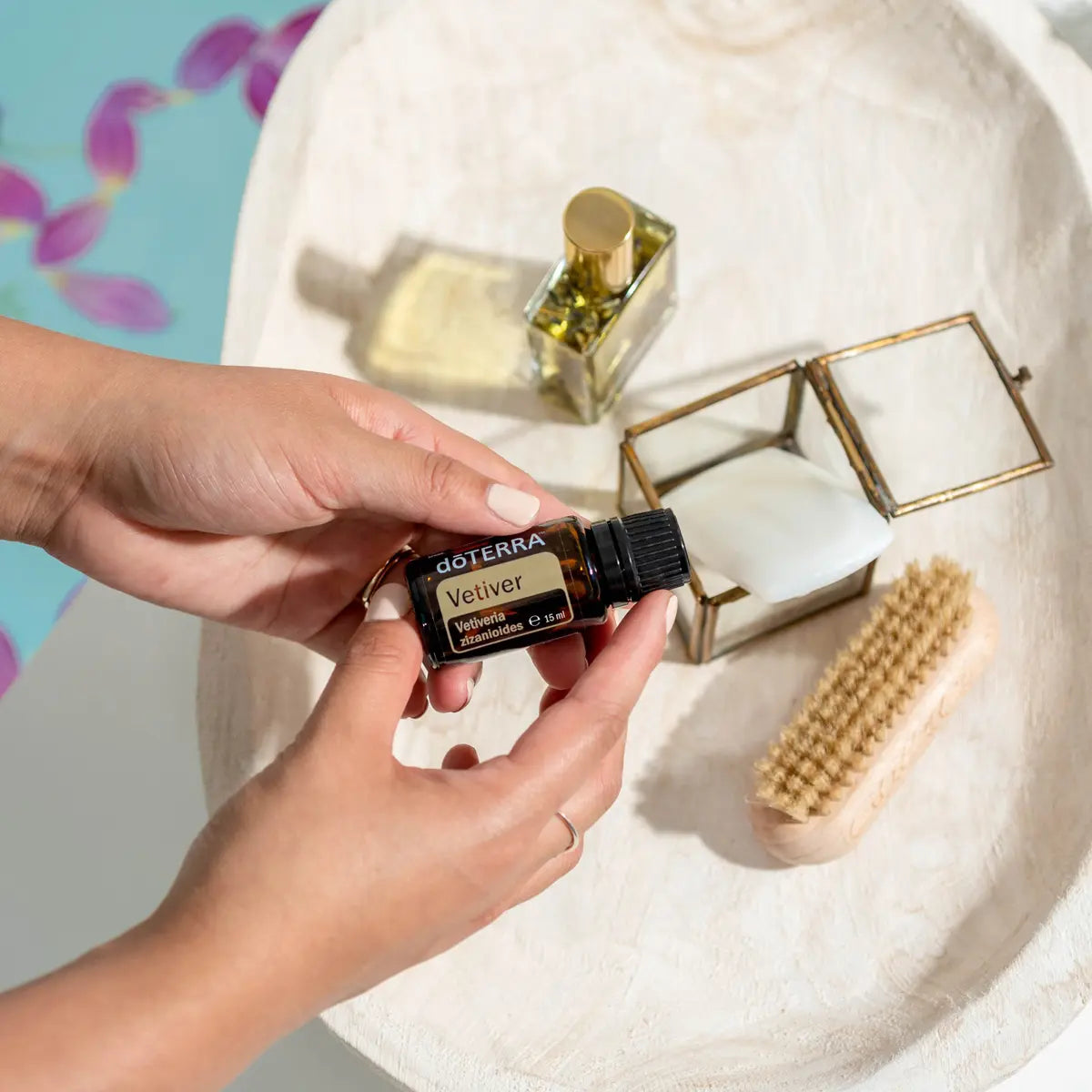 Manos sosteniendo el Aceite Esencial de Vetiver dōTERRA 15ml sobre bandeja de piedra redondeada con frasco de perfume dorado, caja de vidrio con latón y cepillo de uñas de madera, pétalos de orquídea desenfocados