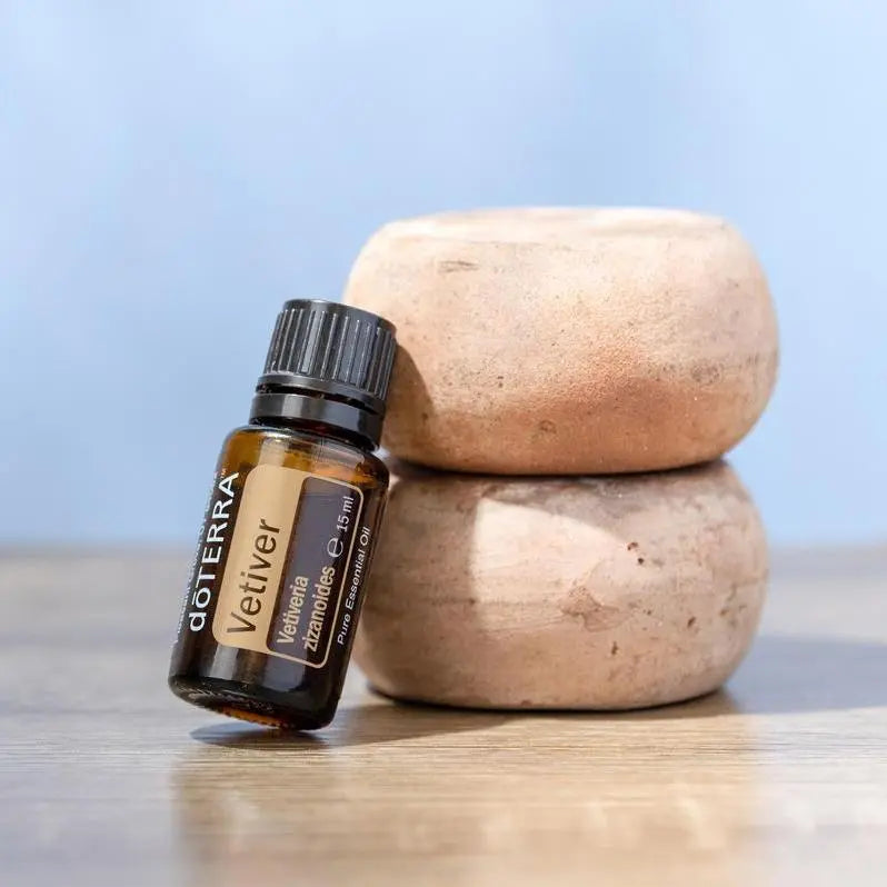 Frasco del Aceite Esencial de Vetiver dōTERRA 15ml apoyado contra dos piedras arenisca apiladas sobre superficie de madera, fondo azul claro