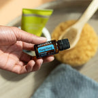 Mano sosteniendo el Aceite Esencial de Ylang Ylang dōTERRA 15ml con cuchara de madera con scrub de azúcar y tubo verde desenfocados al fondo, ambiente de spa