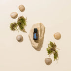 Frasco del Aceite Esencial de Ylang Ylang dōTERRA 15ml sobre piedra plana rústica rodeado de guijarros redondeados y musgo verde disperso, fondo crema