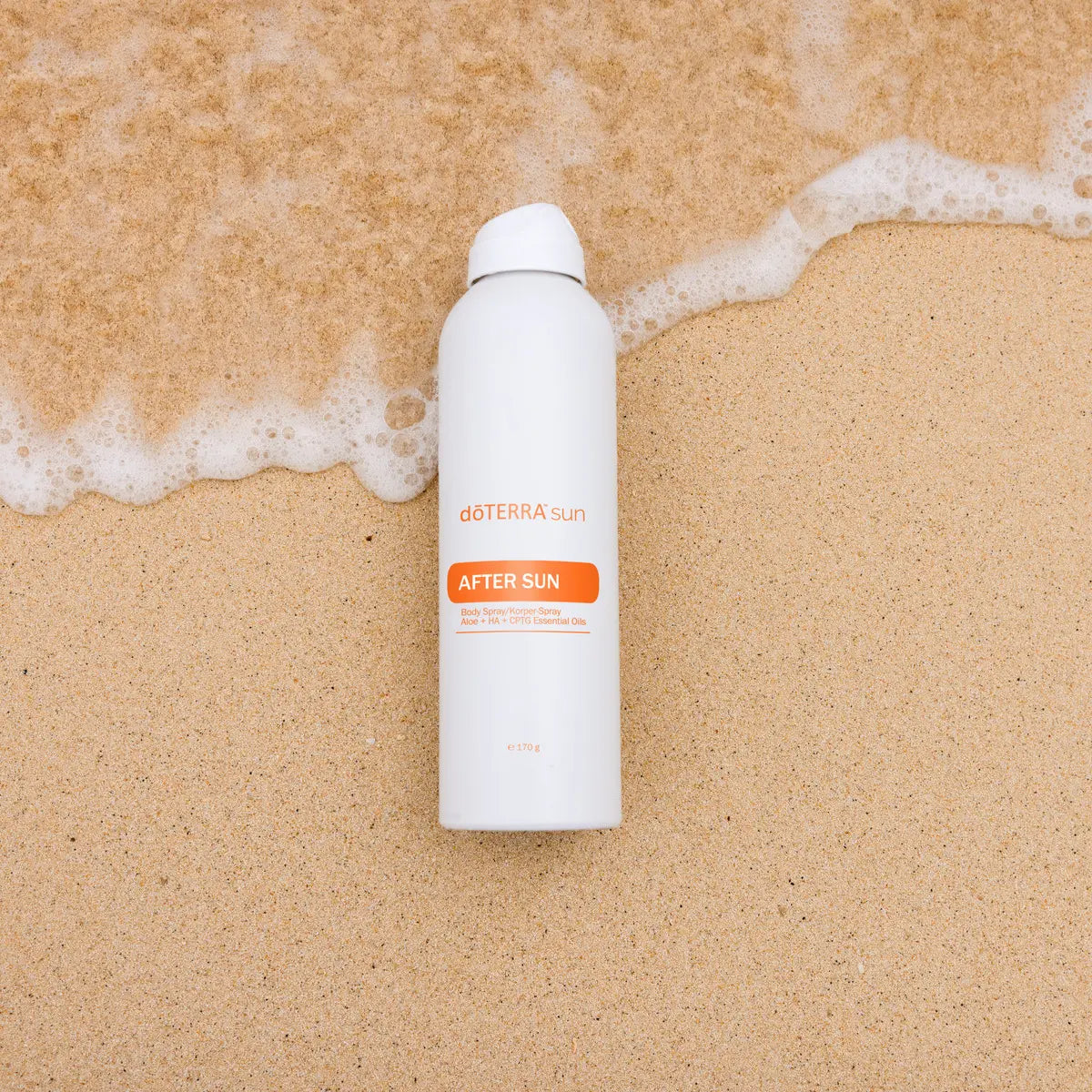 After Sun dōTERRA spray 170g — frasco en arena húmeda con espuma de ola, vista cenital