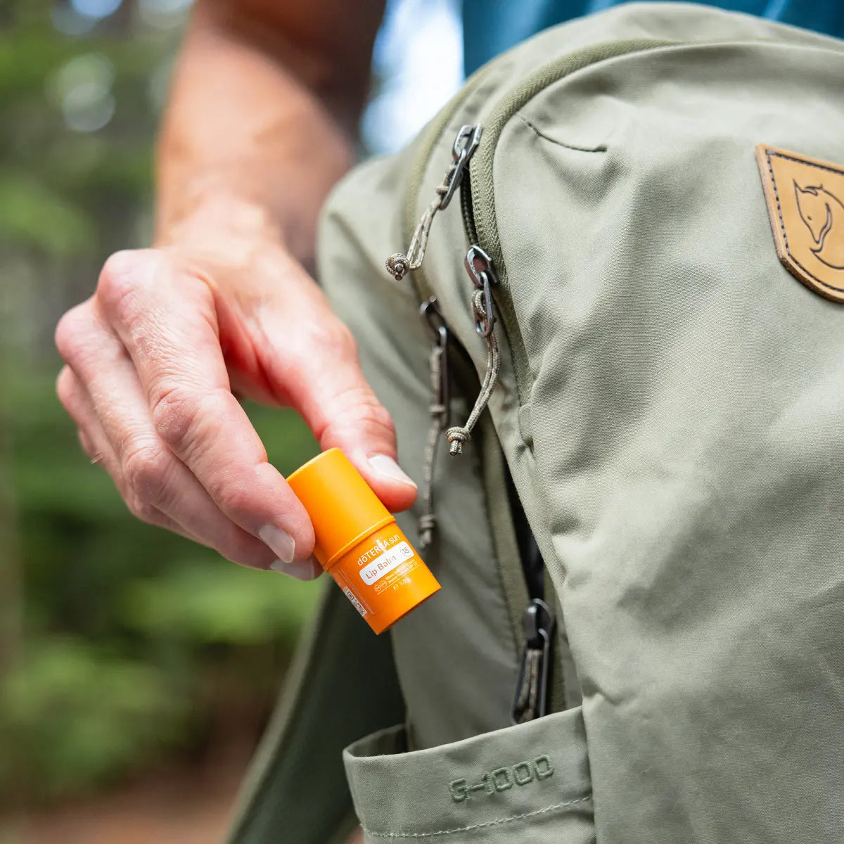 Bálsamo labial Sun dōTERRA SPF 15 — mano sacando del bolsillo de mochila de trekking en bosque