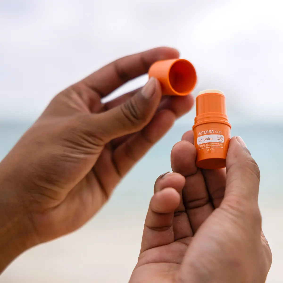 Bálsamo labial Sun dōTERRA SPF 15 — manos abriendo el frasco en la playa con fondo de mar