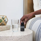 Mano alcanzando el frasco de Capsulas Blandas CP+ doTERRA sobre mesita terrazzo junto a vaso de agua, sofa de lino rayado y terrario de cristal al fondo