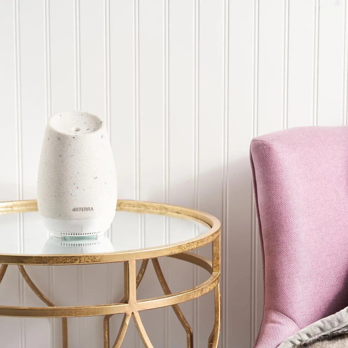 Difusor Roam dōTERRA sobre mesa de cristal dorada junto a silla rosa en el salón