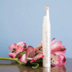 Aplicador en tubo de la crema para contorno de ojos dōTERRA Anti-Aging Eye Cream con esfera de masaje, en pie junto a flores rosas sobre fondo azul claro.