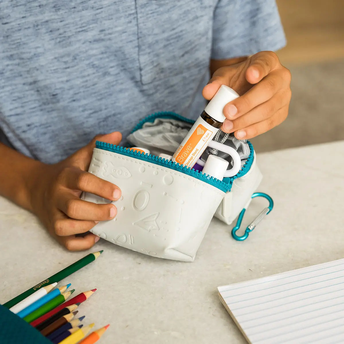 Niño sacando el roll-on dōTERRA Brave Touch de 10ml de un estuche blanco junto a lápices de colores y libreta.