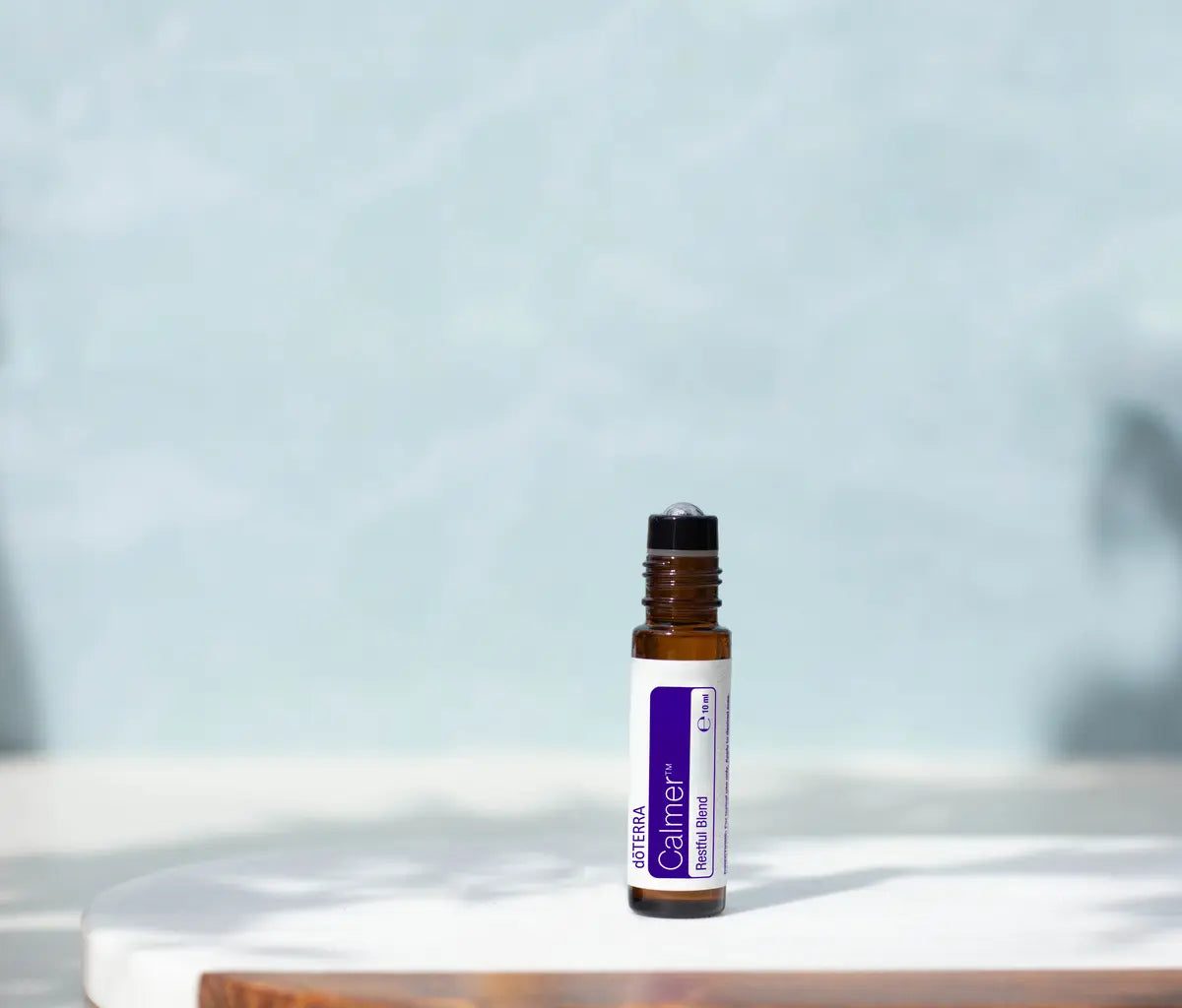 Frasco roll-on dōTERRA Calmer Touch 10ml destapado sobre bandeja de mármol con sombras de hojas y fondo azul suave.