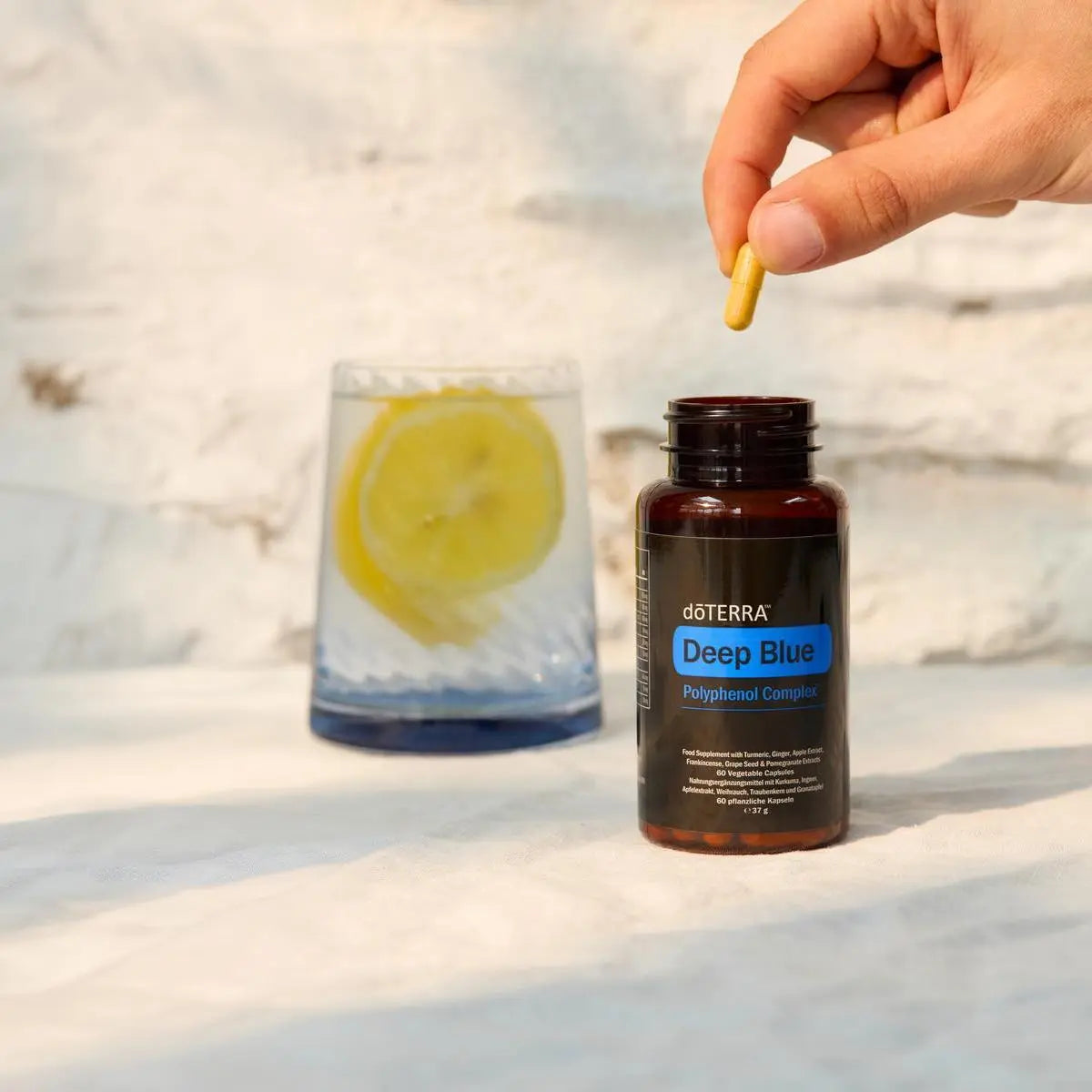 Mano sosteniendo cápsula doTERRA Deep Blue Polyphenol Complex junto a vaso de agua con limón y hielo
