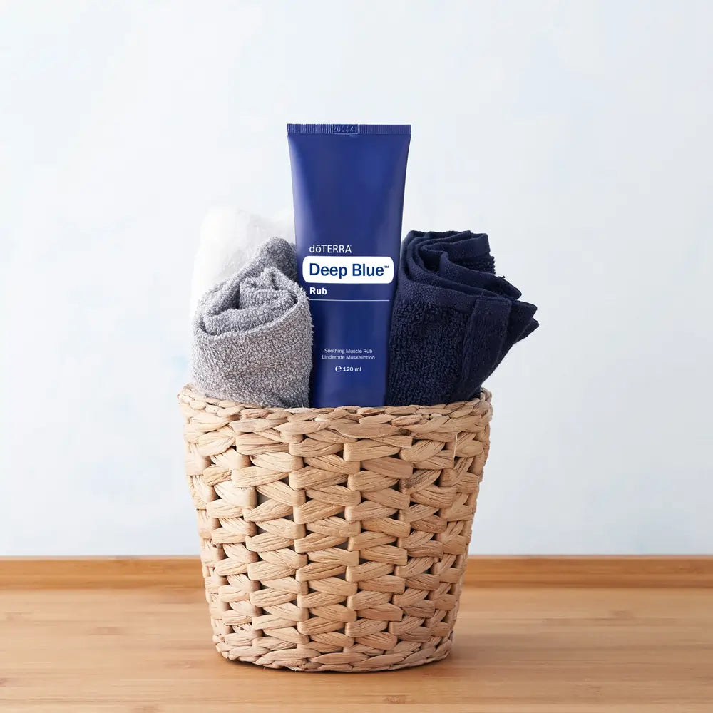 Tubo azul de crema muscular dōTERRA Deep Blue Rub 120ml dentro de una cesta de mimbre con toallas dobladas sobre encimera de madera.