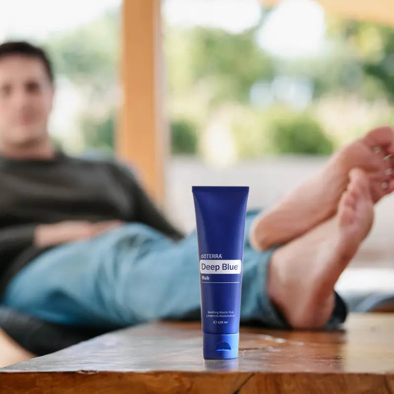 Tubo de crema muscular dōTERRA Deep Blue Rub 120ml en primer plano con hombre relajado con pies descalzos al fondo.