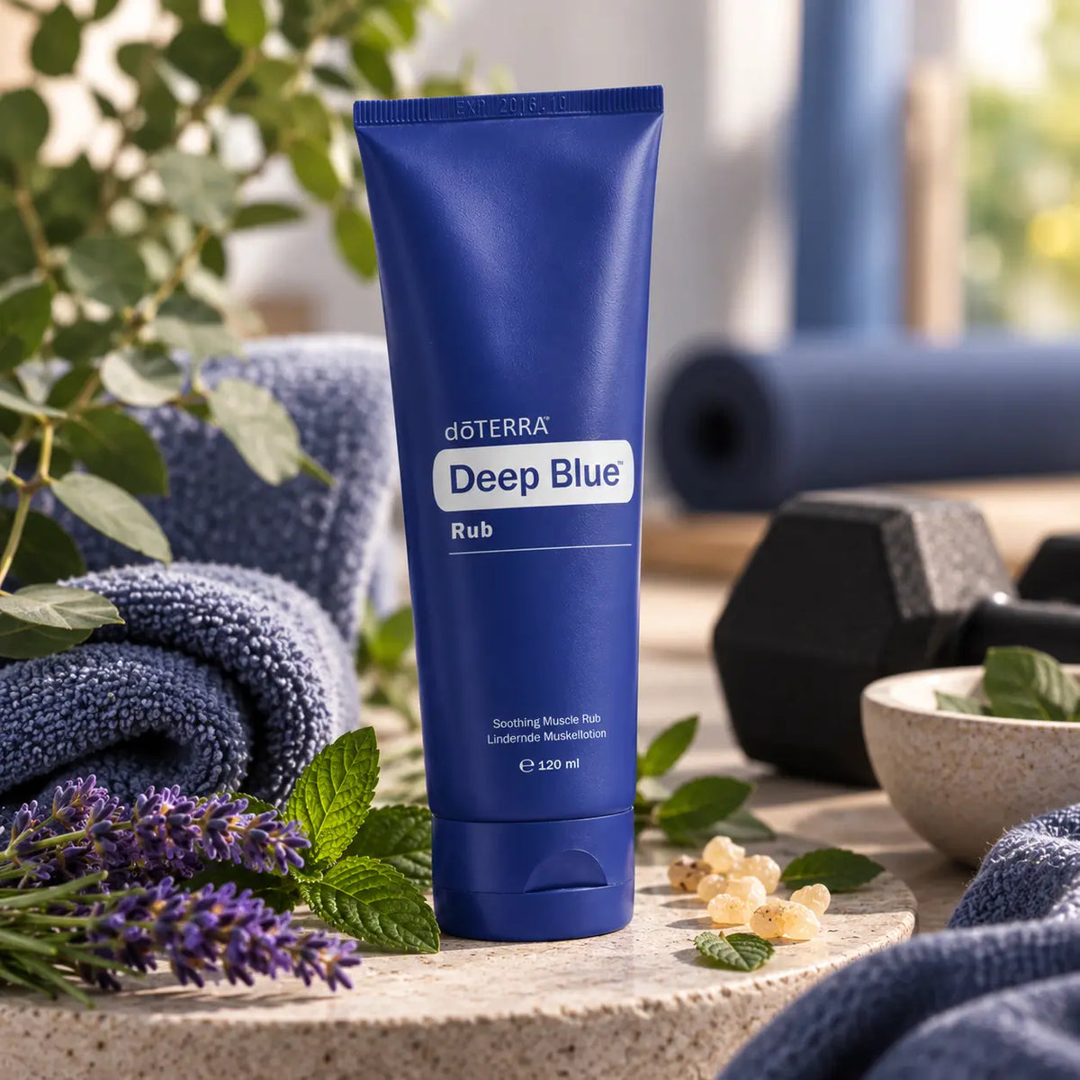 Tubo de crema muscular dōTERRA Deep Blue Rub 120ml sobre piedra con ramos de lavanda, menta, resina de incienso y mancuerna al fondo.