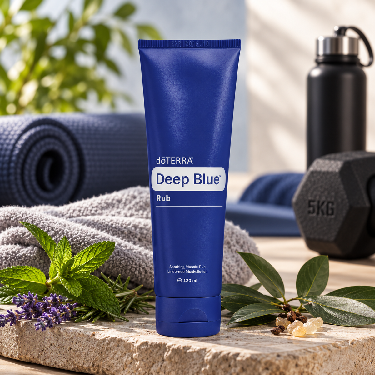 Tubo azul de crema muscular dōTERRA Deep Blue Rub 120ml rodeado por hojas de menta, lavanda, mancuerna de 5kg y esterilla de entrenamiento.