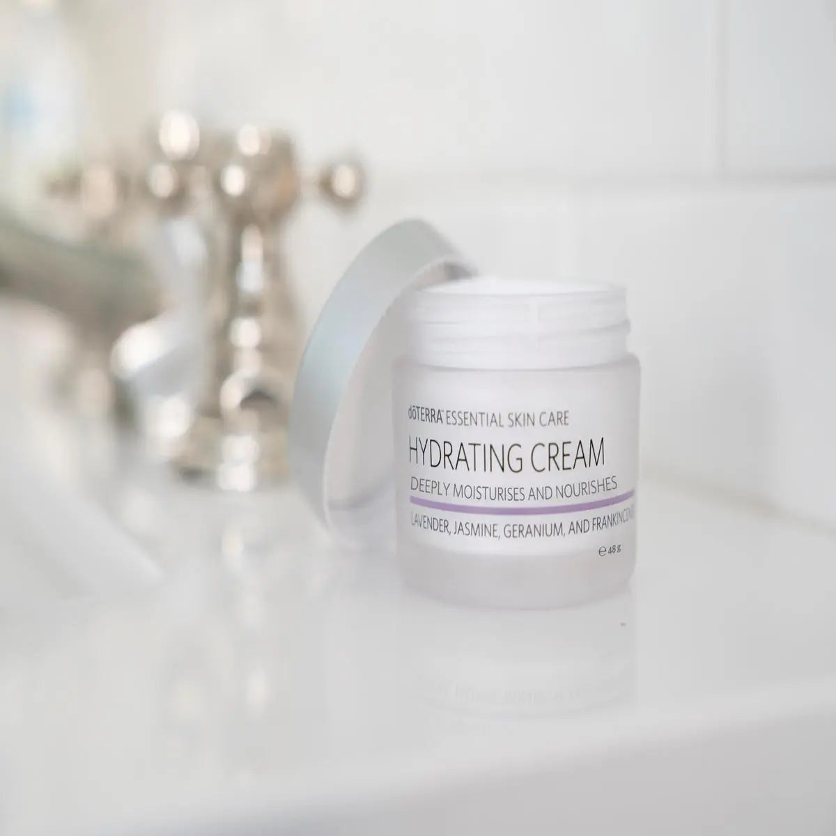 Tarro abierto de dōTERRA Hydrating Cream 48g con la tapa apoyada sobre la encimera de un lavabo de baño.