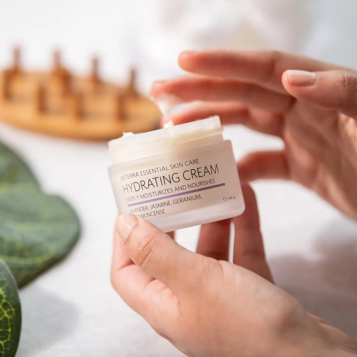 Manos sosteniendo el tarro abierto de dōTERRA Hydrating Cream 48g y tomando crema con la yema de los dedos.