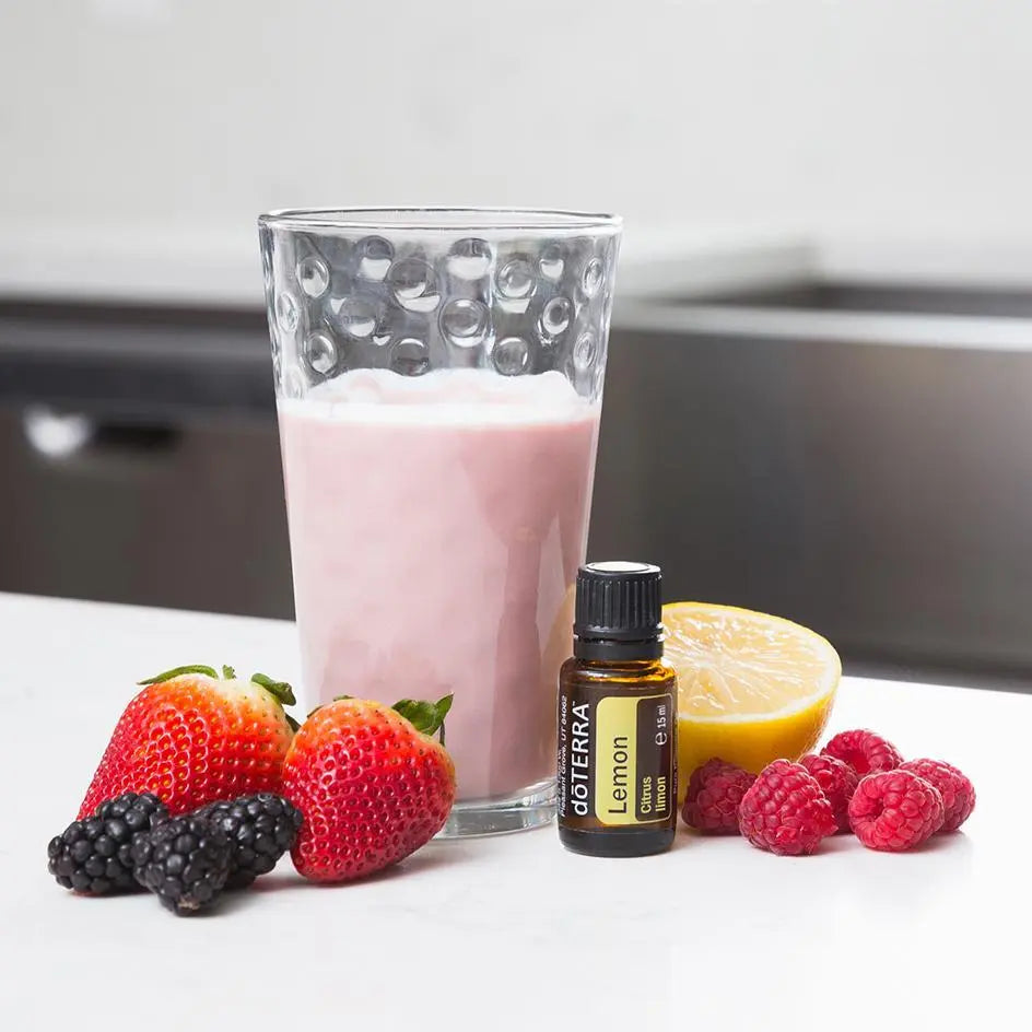 Frasco de dōTERRA Lemon 15ml frente a un vaso de batido rosa rodeado por fresas, frambuesas, moras y medio limón.