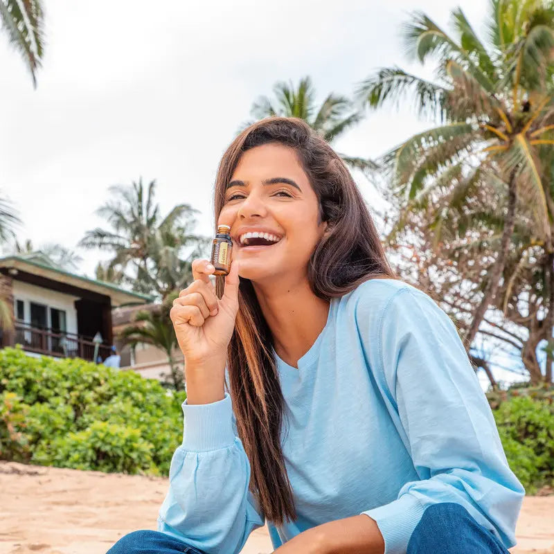 Mujer sonriente sentada en la playa sosteniendo un frasco de aceite esencial dōTERRA Lemon 15ml junto al rostro.